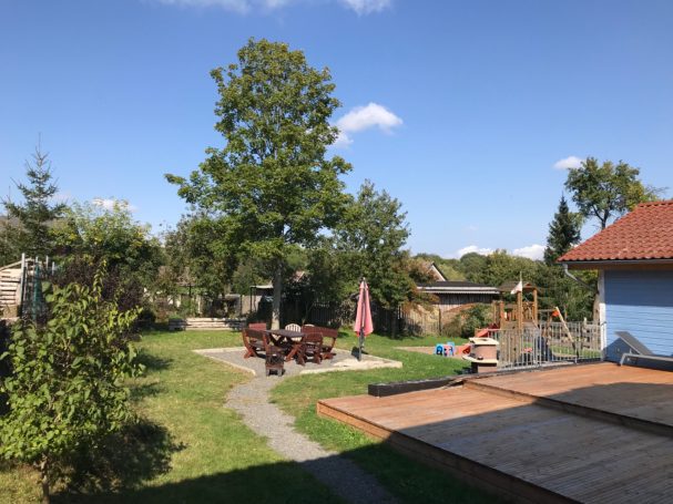 Grüner Garten mit Sitzbereich, Bäumen und einem Weg unter blauem Himmel.